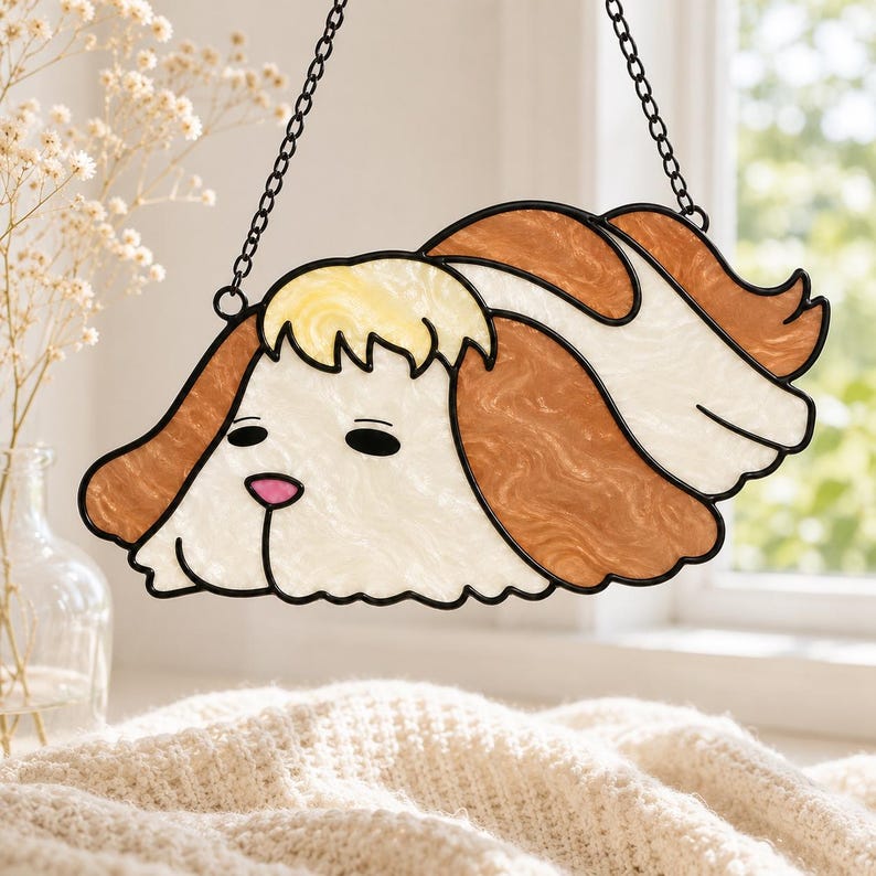 May include: A stained glass suncatcher shaped like a dog head with long, wavy brown and white ears, a pale yellow tuft of hair on the forehead, and a small pink nose. The piece is outlined in black metal and hangs from a thin black metal chain.