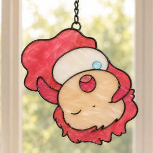 May include: Stained glass suncatcher featuring a whimsical face with a red hat and hair, a white cap, and a light blue gem. The face is a mix of peach and cream colors, outlined in black, and hangs from a chain.