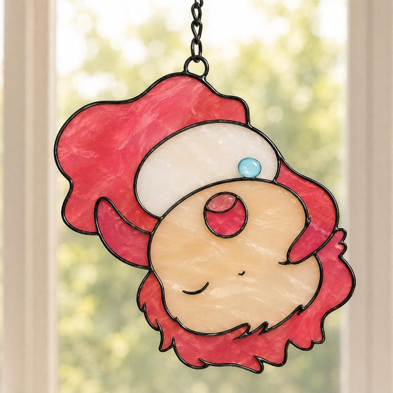May include: Stained glass suncatcher featuring a whimsical face with a red hat and hair, a white cap, and a light blue gem. The face is a mix of peach and cream colors, outlined in black, and hangs from a chain.