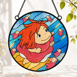 May include: A circular stained glass suncatcher featuring a central character with bright orange hair and a red dress, surrounded by small pink figures against a blue wavy background. The piece is framed in black metal and hangs from a chain.