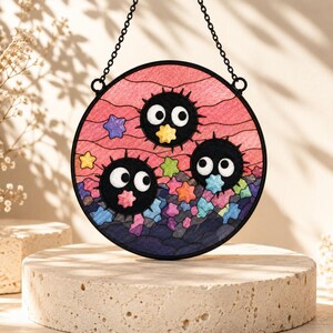 May include: A circular stained glass suncatcher featuring three black, spiky soot sprites with large white eyes. The background displays a pink sky with wavy lines, colorful small stars, and a dark blue rocky base, all framed in black metal.