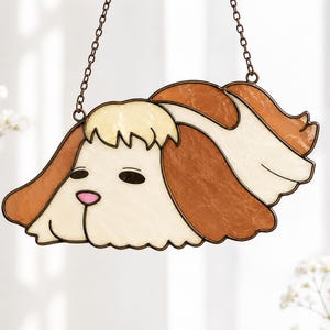 May include: A stained glass suncatcher shaped like a dog head with long, drooping ears. The piece features cream-colored glass for the face and ears, accented with brown glass sections and a small pink glass nose, all held together by dark metal cames.