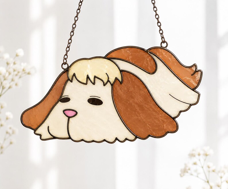 May include: A stained glass suncatcher shaped like a dog head with long, drooping ears. The piece features cream-colored glass for the face and ears, accented with brown glass sections and a small pink glass nose, all held together by dark metal cames.