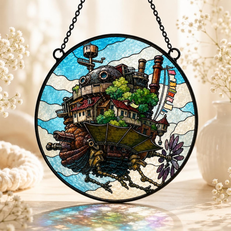 May include: A circular stained glass suncatcher featuring a detailed depiction of a mechanical walking castle with multiple chimneys, green foliage, and a colorful flag banner, suspended by a black metal chain against a blue and white sky background.