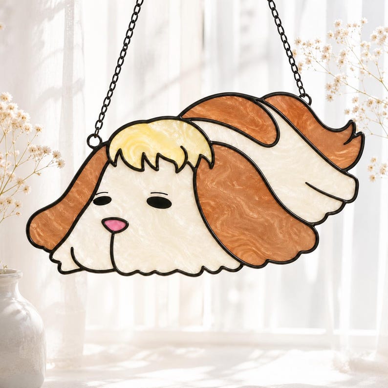 May include: A stained glass suncatcher shaped like a dog head with long, flowing ears. The piece features cream, tan, and brown marbled glass segments outlined in black metal, with a small pink glass nose and two black oval eyes hanging from a chain.