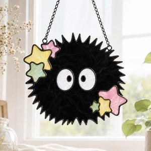 May include: A stained glass suncatcher featuring a round, spiky black creature with two large white eyes. The design is accented with small, pastel-colored stars in shades of pink, yellow, and light green. It hangs from a black metal chain.
