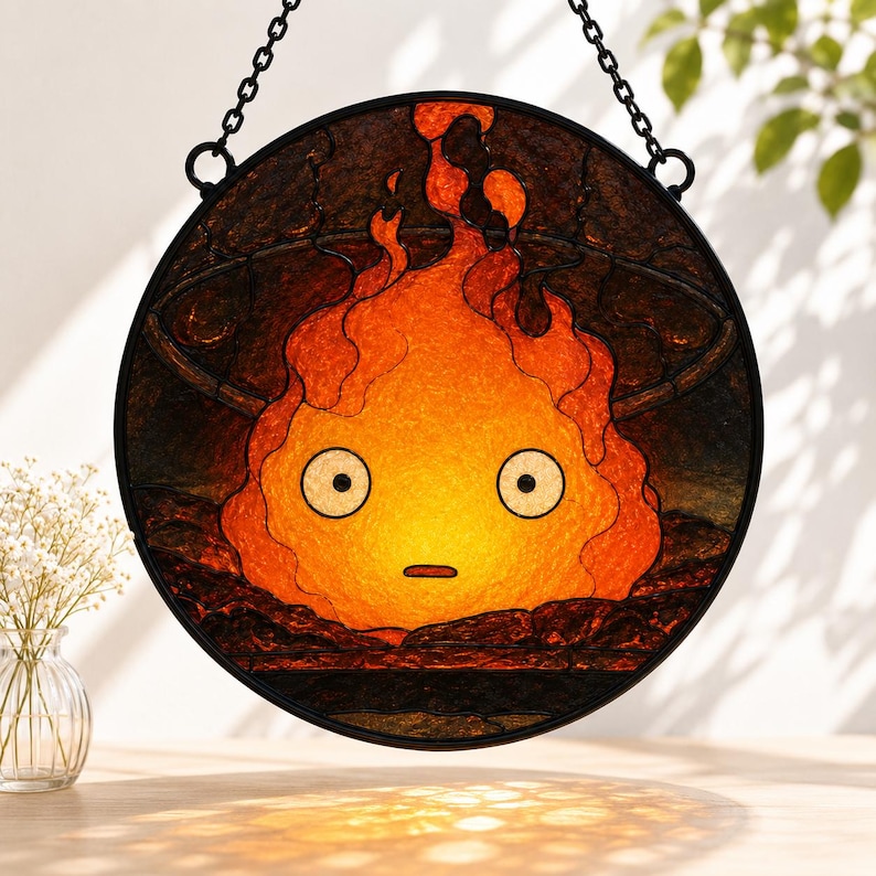 Puede incluir: Un atrapasoles circular de vidrieras que presenta un personaje de llama naranja y amarillo estilizado con grandes ojos blancos y una boca peque&ntilde;a. El dise&ntilde;o est&aacute; encerrado en un marco de metal negro y cuelga de una cadena negra.