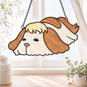 May include: A stained glass suncatcher shaped like a dog with long, wavy brown and white fur, a pale yellow tuft on its head, and a small pink nose. The piece is outlined in black metal and hangs from a thin black chain against a window.
