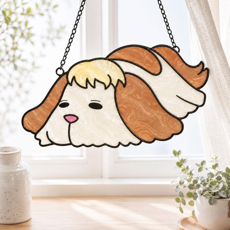 May include: A stained glass suncatcher shaped like a dog with long, wavy brown and white fur, a pale yellow tuft on its head, and a small pink nose. The piece is outlined in black metal and hangs from a thin black chain against a window.
