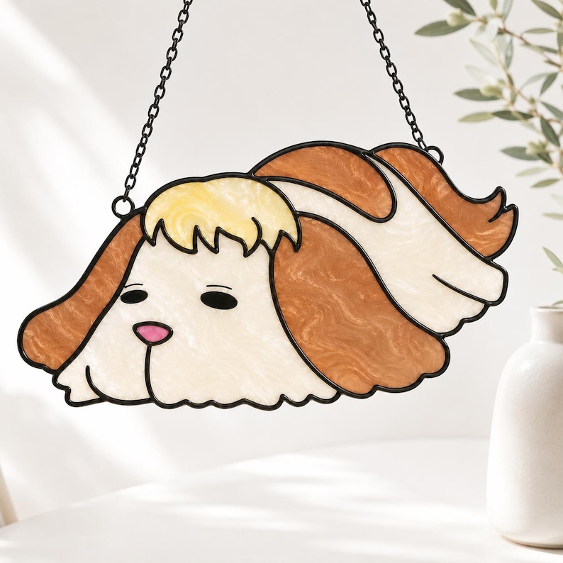 May include: A stained glass suncatcher shaped like a dog head with long, wavy brown and cream-colored ears. The piece features a pale yellow top section, a pink nose, and black glass outlines, suspended by a thin black metal chain for hanging.