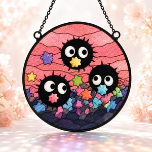 May include: A circular stained glass suncatcher featuring three black, spiky soot sprites with large white eyes. The background displays a pink sky with wavy lines and a cluster of colorful, multi-shaped stars in shades of blue, green, and orange.