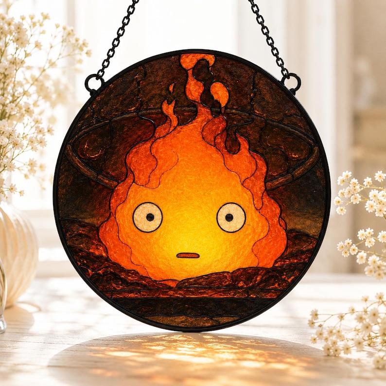 Puede incluir: Un atrapasue&ntilde;os circular de vidrieras que presenta un personaje de llama naranja y amarillo estilizado con grandes ojos blancos y una boca peque&ntilde;a, sobre un fondo oscuro y texturizado, suspendido por una cadena de metal negro y un marco.