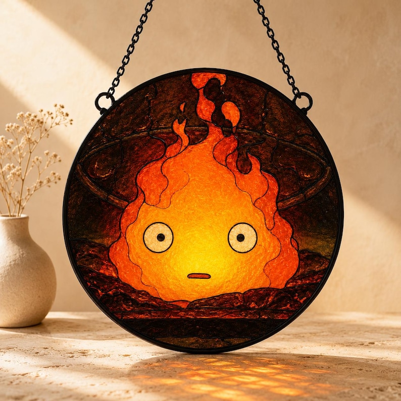 Puede incluir: Un atrapasoles circular de vidriera que presenta un personaje de llama naranja y amarillo estilizado con grandes ojos blancos y una boca peque&ntilde;a, enmarcado en metal negro con una cadena, sobre un fondo oscuro y texturizado.