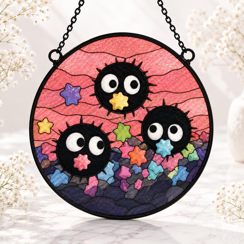 May include: A circular stained glass suncatcher featuring three black, spiky, round soot sprites with large white eyes. The background is a textured pink sky with numerous small, colorful stars in shades of yellow, green, blue, and purple.