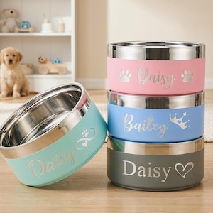 May include: A stack of personalized pet bowls in pink, blue, and gray, with stainless steel interiors. Each bowl features a pet's name and decorative paw prints or a crown. One teal bowl is tilted, displaying the name "Daisy".