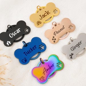 Puede incluir: Una colección de etiquetas para mascotas en forma de hueso, en varios colores, incluyendo negro, dorado, azul, rosa, plateado y arcoíris. Cada etiqueta está personalizada con el nombre de una mascota, como "Oscar", "Jack" y "Amber", junto con huellas de patas e iconos.