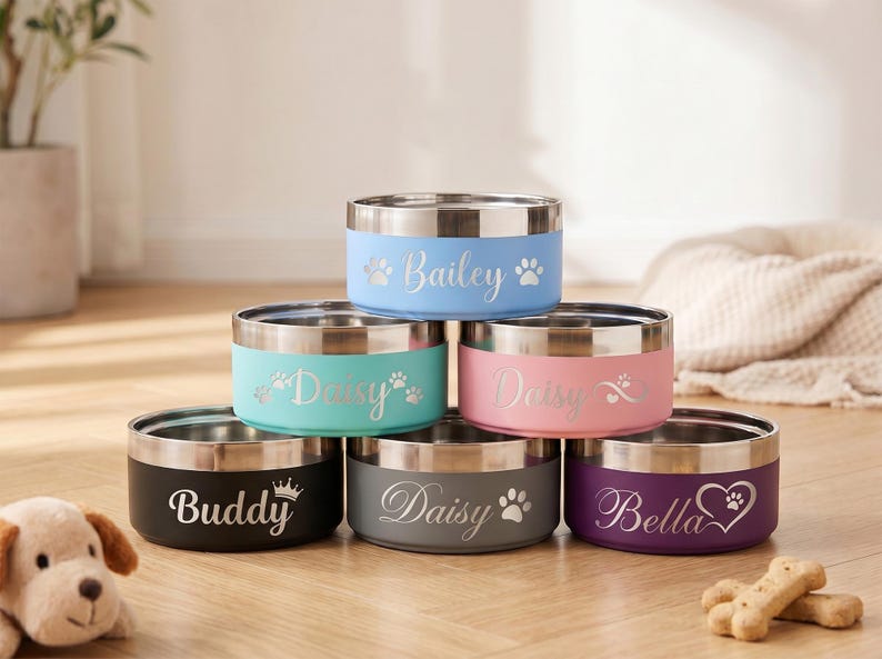 May include: A stack of six stainless steel pet bowls with colored exteriors. Each bowl has a silver rim and is personalized with a pet's name and paw print or heart design. Colors include blue, teal, pink, black, gray, and purple.