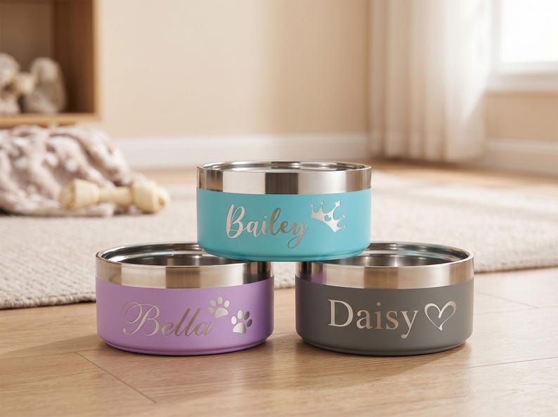 May include: Three stainless steel pet bowls in teal, lavender, and gray. Each bowl has a silver rim and a personalized name: "Bailey" with a crown, "Bella" with paw prints, and "Daisy" with a heart.