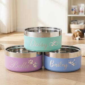 May include: Three stainless steel pet bowls in pastel colors: mint, lavender, and blue. Each bowl has a silver rim and is personalized with a pet's name in script. The mint bowl says "Daisy", the lavender bowl says "Bella", and the blue bowl says "Bailey".