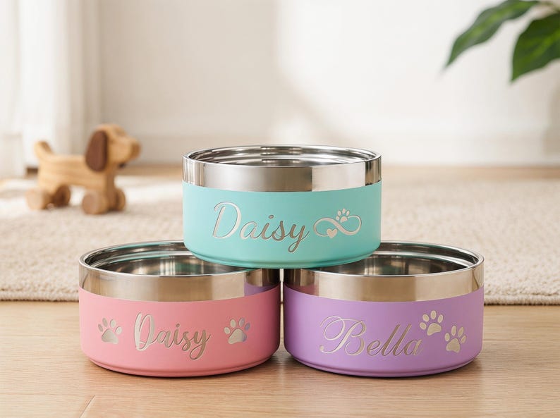 May include: Three stainless steel pet bowls in pastel colors: light blue, pink, and purple. Each bowl has a silver rim and is personalized with a pet's name in cursive script. Paw prints and hearts are also featured.