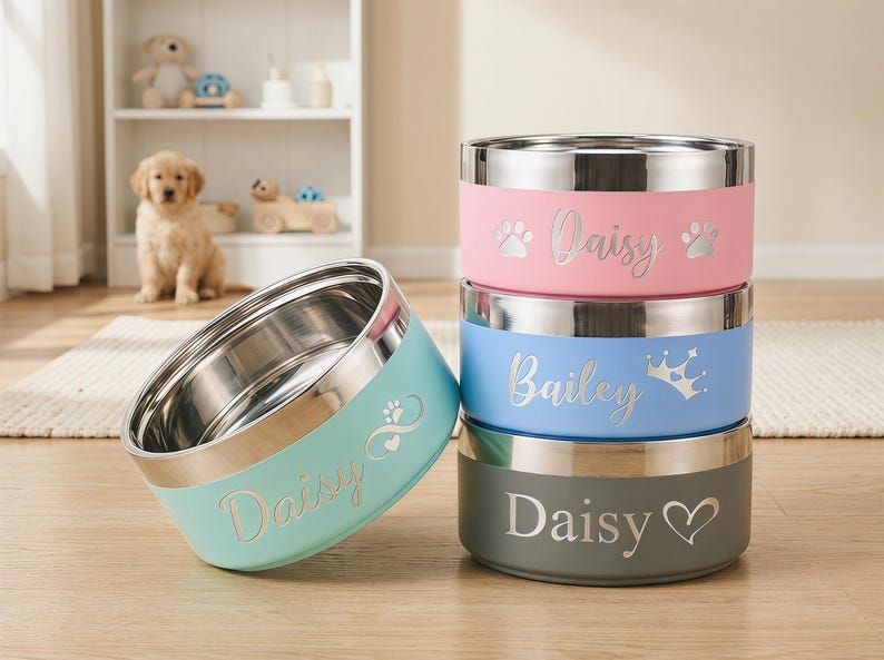 May include: A stack of personalized pet bowls in pink, blue, and gray, with stainless steel interiors. Each bowl features a pet's name and decorative paw prints or a crown. One teal bowl is tilted, displaying the name "Daisy".