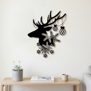 May include: A black metal wall art piece shaped like a deer head in profile. The deer antlers are adorned with two hanging spherical ornaments, and the neck area is decorated with several intricate, layered snowflake cutouts in black metal.