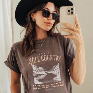 K&ouml;nnte beinhalten: Eine Frau tr&auml;gt ein dunkelbraunes T-Shirt mit hochgekrempelten &Auml;rmeln, Sonnenbrille und einen schwarzen Hut. Das Shirt zeigt den Text "ONLY IN HILL COUNTRY AS IT IS IN HEAVEN" mit einer Landschaftsgrafik.