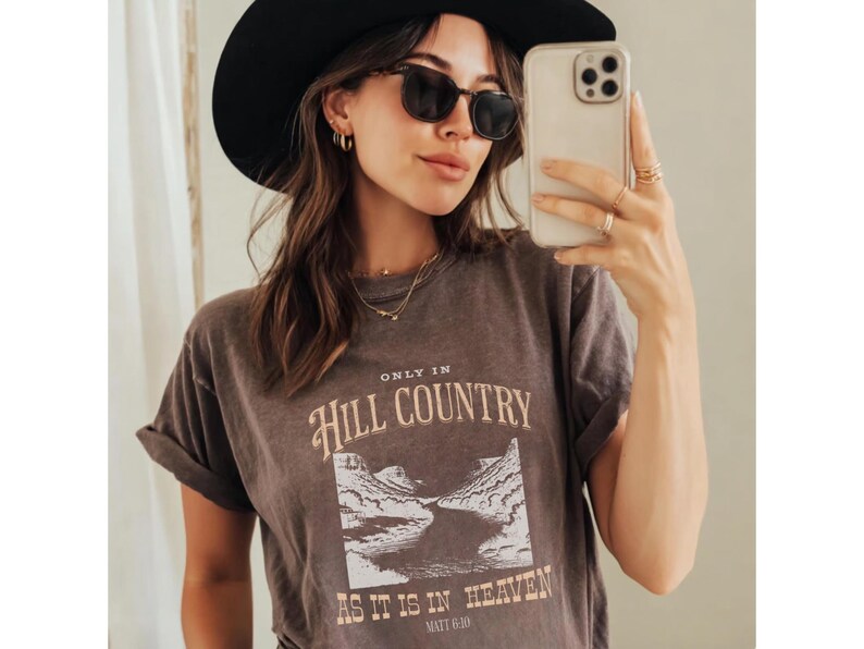 K&ouml;nnte beinhalten: Eine Frau tr&auml;gt ein dunkelbraunes T-Shirt mit hochgekrempelten &Auml;rmeln, Sonnenbrille und einen schwarzen Hut. Das Shirt zeigt den Text "ONLY IN HILL COUNTRY AS IT IS IN HEAVEN" mit einer Landschaftsgrafik.
