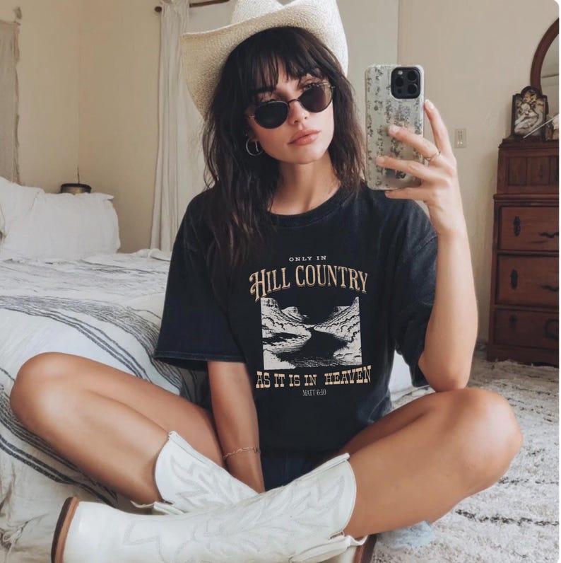 K&ouml;nnte beinhalten: Schwarzes T-Shirt mit dem Text "ONLY IN HILL COUNTRY AS IT IS IN HEAVEN" und einer Landschaftsgrafik. Die Person tr&auml;gt einen wei&szlig;en Cowboyhut, eine Sonnenbrille und wei&szlig;e Cowboy-Stiefel.