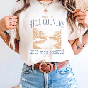 K&ouml;nnte beinhalten: Cremefarbenes T-Shirt mit dem Text "ONLY IN Hill COUNTRY AS IT IS IN HEAVEN" in Blau und Beige. Das Design beinhaltet eine Landschaftsillustration. Das Shirt wird mit Jeansshorts und einem braunen G&uuml;rtel getragen.