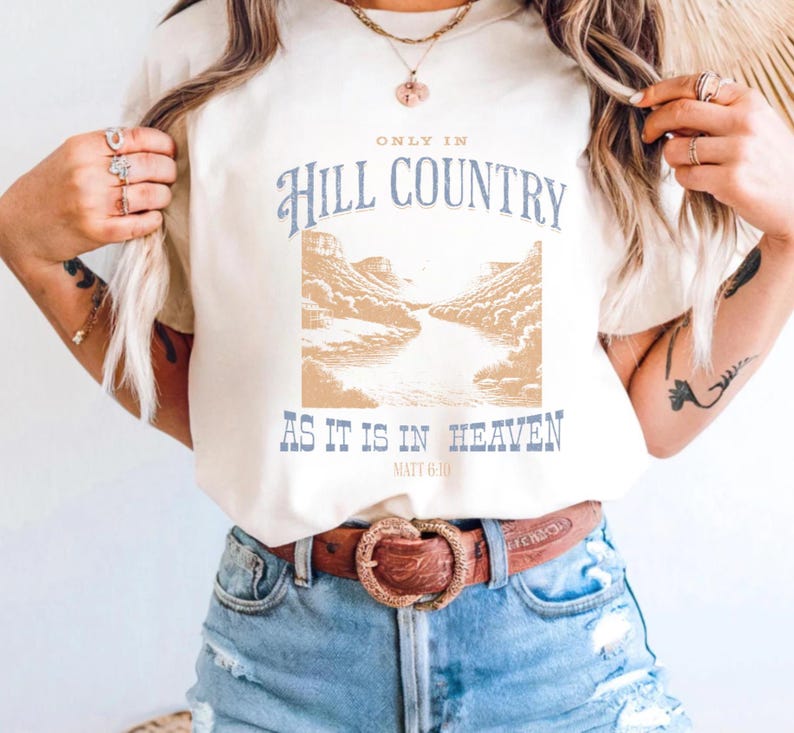 K&ouml;nnte beinhalten: Cremefarbenes T-Shirt mit dem Text "ONLY IN Hill COUNTRY AS IT IS IN HEAVEN" in Blau und Beige. Das Design beinhaltet eine Landschaftsillustration. Das Shirt wird mit Jeansshorts und einem braunen G&uuml;rtel getragen.