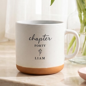 以下が含まれることがあります： 茶色の底と「chapter forty」と「LIAM」の文字が入った白いセラミックマグ。単語の間に小さな花のイラストがあります。マグカップは大理石の表面にあり、背景には花の入った花瓶があります。