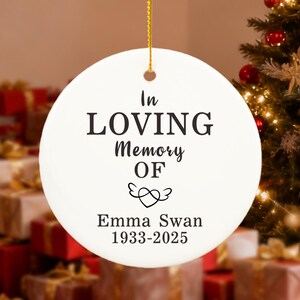 Può includere: Un ornamento natalizio rotondo bianco con il testo nero "In Loving Memory Of Emma Swan 1933-2025". È appeso a un cordino dorato. Un albero di Natale e regali confezionati sono sullo sfondo.