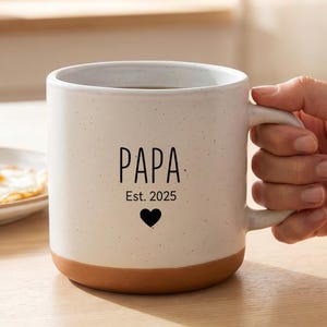 Könnte beinhalten: Eine weiße Keramiktasse mit braunem Boden, mit dem Wort "PAPA", dem Jahr "Est. 2025" und einem schwarzen Herzen. Die Tasse wird von einer Person gehalten. Ein Teller mit Spiegelei und Toast befindet sich im Hintergrund.