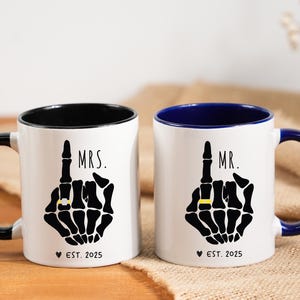 May include: Two white ceramic mugs with black and blue accents. Each mug features a black skeleton hand with a gold ring, the word "MRS." or "MR.", and "EST. 2025". The mugs are on a wooden surface.