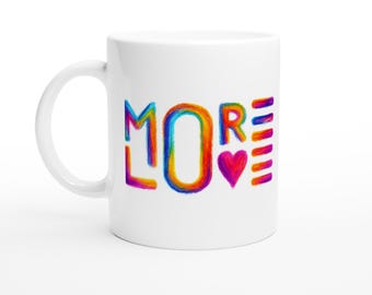 Può includere: Tazza in ceramica bianca con la scritta "MORE" in lettering arcobaleno, stile scritto a mano. Un cuore rosso e strisce verticali completano il design. La tazza ha un manico curvo.