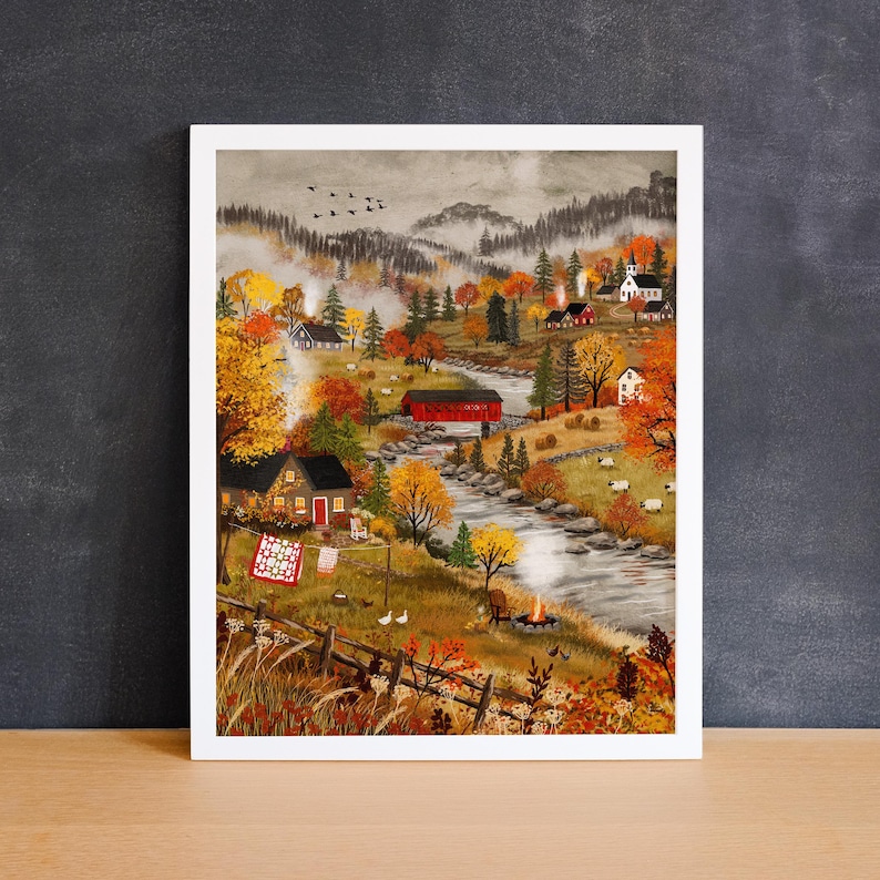 May include: A framed landscape painting depicting a rural autumn scene with a winding river, a red covered bridge, scattered houses, and sheep grazing in fields. The artwork features vibrant orange and yellow trees, a campfire, and a clothesline.