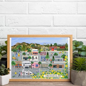May include: A framed art print of Louisville, CO, featuring colorful buildings, trees, and people. The artwork has a light brown wooden frame and is surrounded by potted plants. The text "LOUISVILLE.CO" is visible.