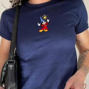 Può includere: Una t-shirt blu navy a maniche corte con un piccolo ricamo raffigurante Topolino in veste di mago con cappello blu e tunica rossa, che tiene in mano una bacchetta a forma di stella dorata. La maglia è abbinata a una borsa a spalla nera.