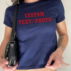 Può includere: T-shirt blu navy con la scritta rossa "CUSTOM TEXT/PHOTO". La maglietta ha maniche corte e girocollo. Una borsa a tracolla nera è visibile sul lato sinistro.