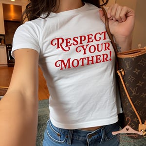 Op de afbeelding: Wit T-shirt met de rode tekst "RESPECT YOUR MOTHER!". Het shirt wordt gecombineerd met een blauwe jeans. Een bruine handtas met monogrampatroon is zichtbaar.