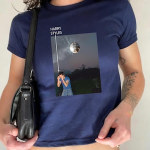 May include: Navy blue t-shirt featuring a graphic of a disco ball and a person in a field. The text "HARRY STYLES" is above the graphic. The shirt has short sleeves.