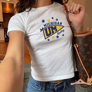 Pode incluir: Uma t-shirt branca de manga curta com um gráfico bordado de uma flâmula amarela e azul com o texto MICHIGAN UM e Hadley, rodeado por pequenas estrelas azuis e amarelas. A t-shirt é combinada com calças de ganga azuis e uma mala castanha.