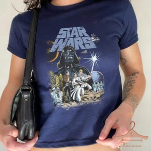 Peut inclure: T-shirt bleu marine Star Wars avec un motif de Dark Vador et d'autres personnages du film. Le graphisme comprend le logo Star Wars et diverses images. Le t-shirt est à manches courtes et a un col rond.