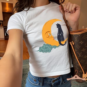May include: White short-sleeved t-shirt with a crescent moon design, a black cat, and a cloud. The graphic is in orange, blue, and black. The shirt is worn with blue jeans. The design has a whimsical feel.