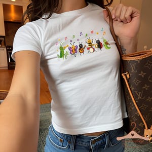 Könnte beinhalten: Ein weißes Kurzarm-T-Shirt mit einem farbenfrohen Grafikdruck von sechs Cartoon-Insekten, die verschiedene Musikinstrumente spielen, darunter eine Heuschrecke, ein Schmetterling, ein Käfer, eine Biene, ein Marienkäfer und eine Raupe.