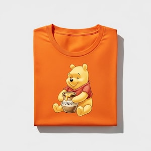 May include: An orange t-shirt featuring a cartoon illustration of Winnie the Pooh holding a honey pot. The honey pot is brown with the word "HUNNY" printed on it. Winnie the Pooh is wearing a red shirt.