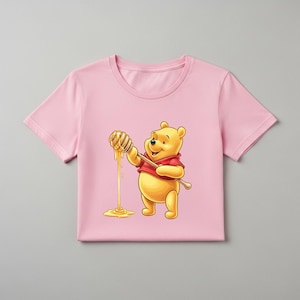 May include: A light pink t-shirt featuring a cartoon illustration of Winnie the Pooh holding a honey dipper with dripping honey. Pooh is wearing a red shirt. The honey is yellow.