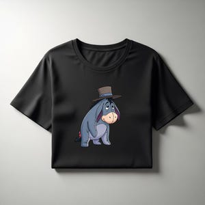 May include: Black t-shirt featuring a cartoon illustration of Eeyore from Winnie the Pooh. Eeyore is depicted in a seated position, wearing a brown hat. The design showcases Eeyore's blue-grey body, pink accents, and a sad expression.