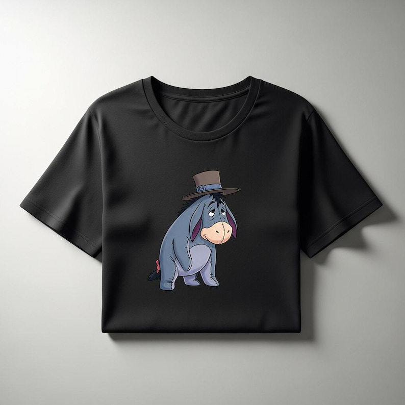 May include: Black t-shirt featuring a cartoon illustration of Eeyore from Winnie the Pooh. Eeyore is depicted in a seated position, wearing a brown hat. The design showcases Eeyore's blue-grey body, pink accents, and a sad expression.